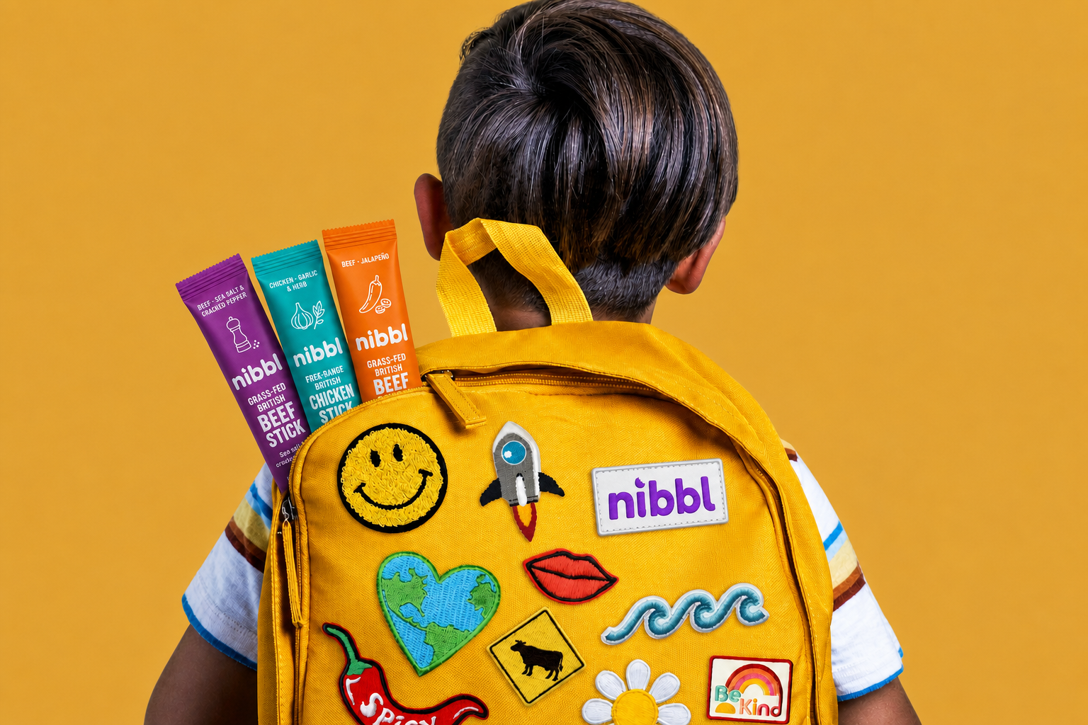 Three Nibbl protein sticks tucked into a kid's yellow backpack covered in patches